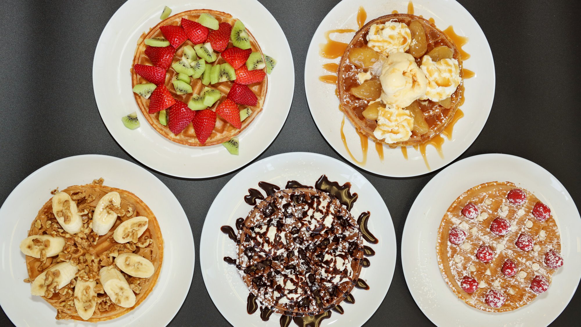 Five Belgian waffles seen from above on a dark table, topped with fruit, whipped cream, chocolate and ice cream